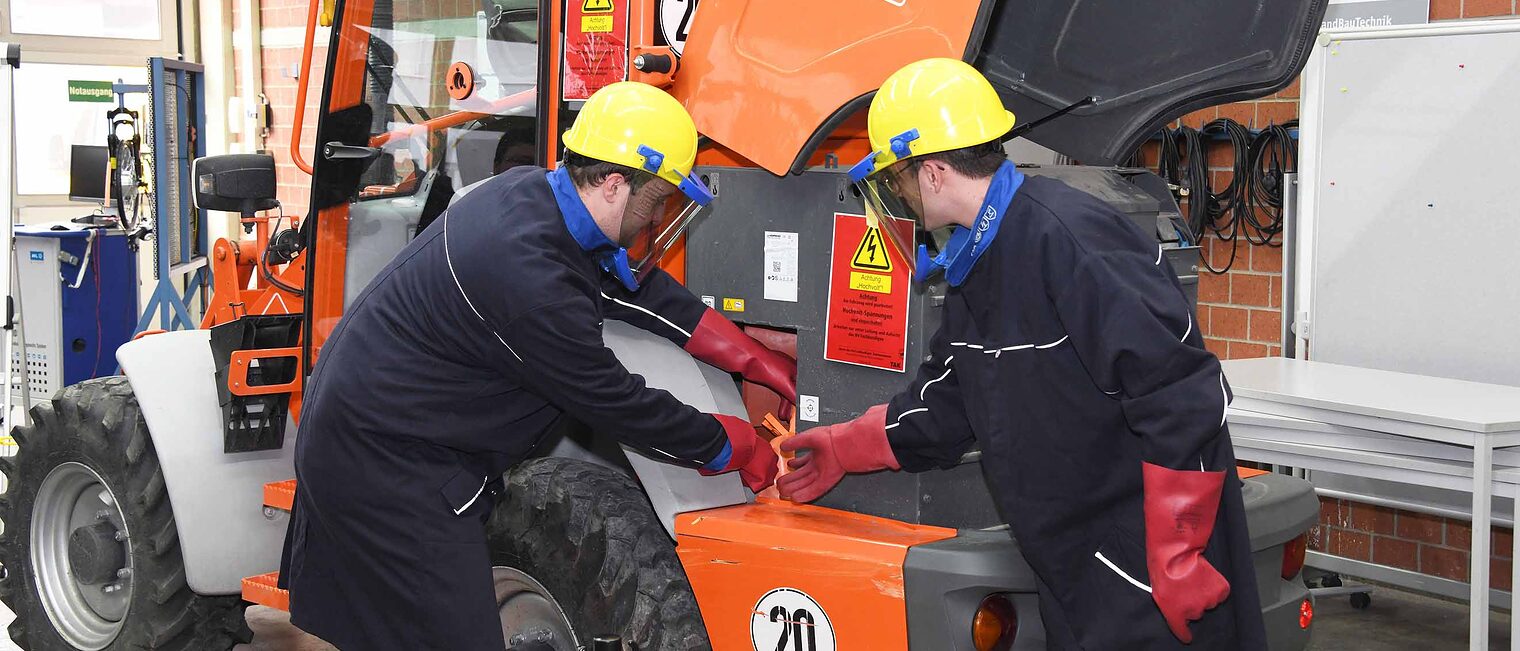 Zwei Männer mit Schutzkleidung führen Arbeiten im Hochvoltbereich an einem orangefarbenen Nutzfahrzeug in der Werkstatt durch