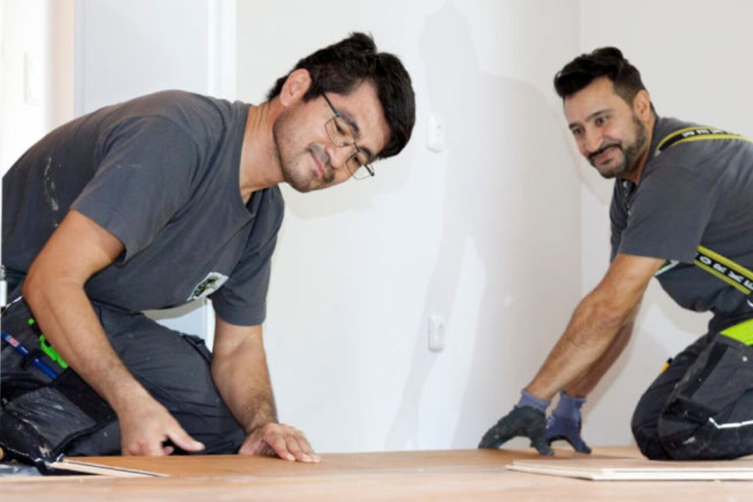 Zwei Männer knien in Arbeitskleidung auf dem Boden und verlegen Parkett