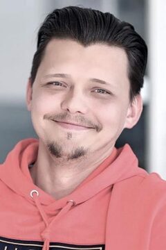 junger Mann, Lächelt, Kapuzenpullover, leichter Kinn- und Schnauzbart Lächelnder junger Mann in hellrotem Kapuzenpullover mit Kinnbart und leichtem Schnauzbart