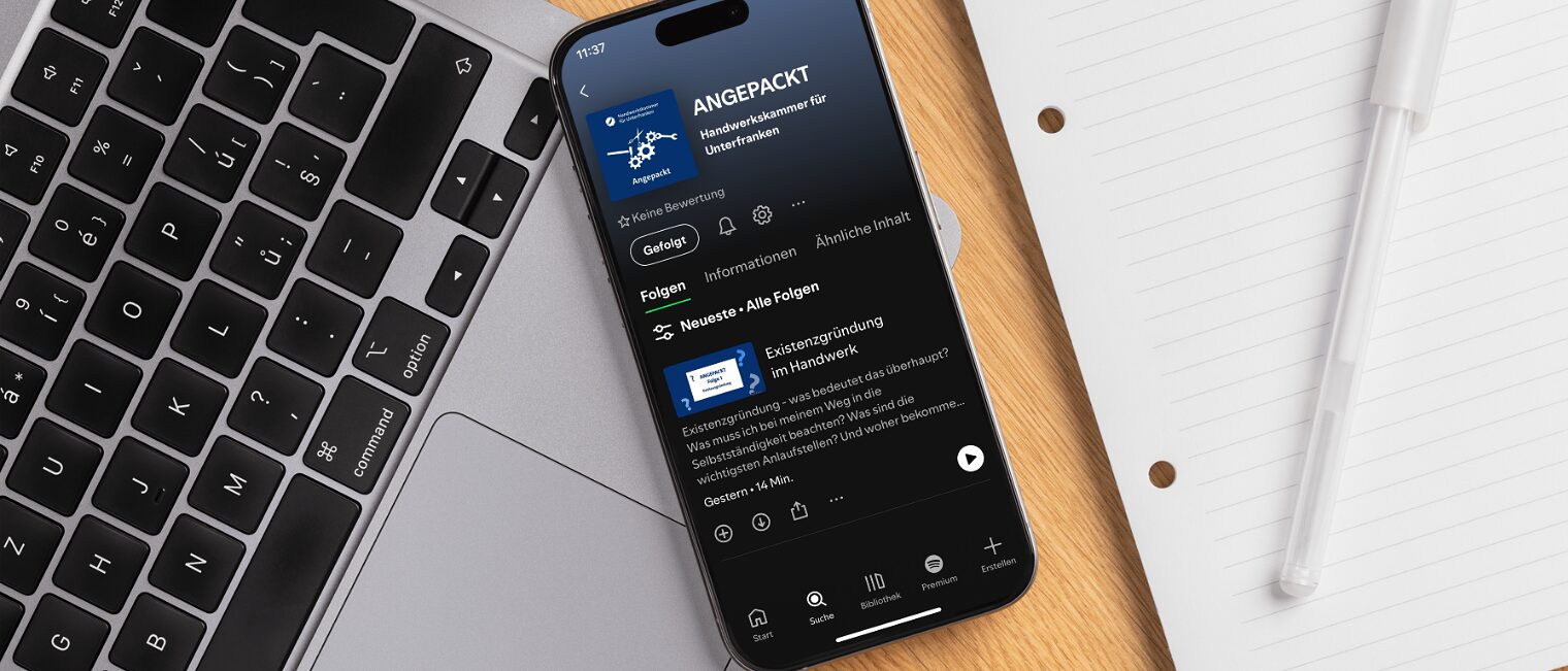 Smartphone mit Bildschirm, auf dem der Podcast "Angepackt" in Spotify geöffnet zu sehen ist, auf einem Schreibtisch.