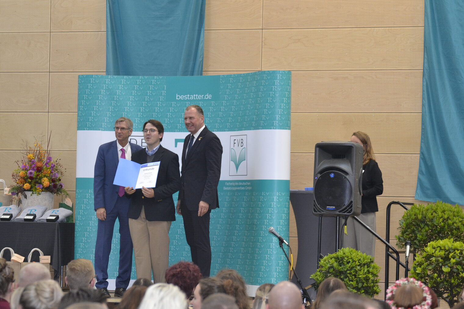 Ehrung einer Person zur Bestattungsfachkraft Drei Menschen sind zu sehen. Sie stehen auf einem Pult und einer bekommt eine Ehrung