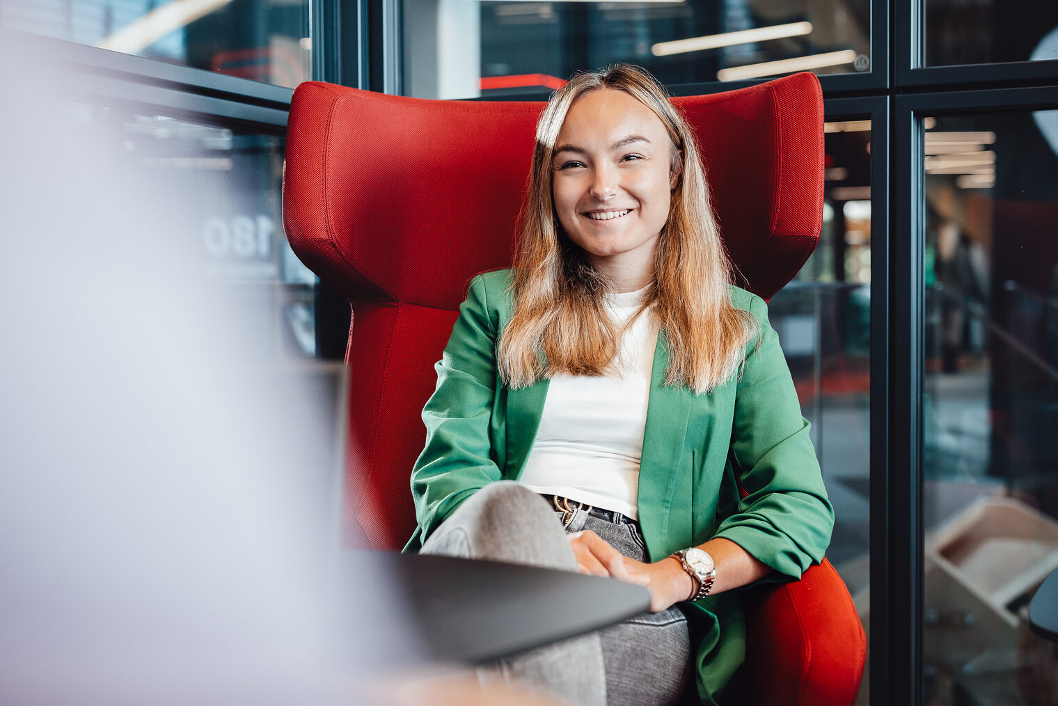 Frau lächelt in die Kamera, trägt grünes Jackett, auf rotem Sessel Frau in grünem Jackett mit mittellangen hellbraunen Haaren sitzt auf rotem Sessel, lächelt