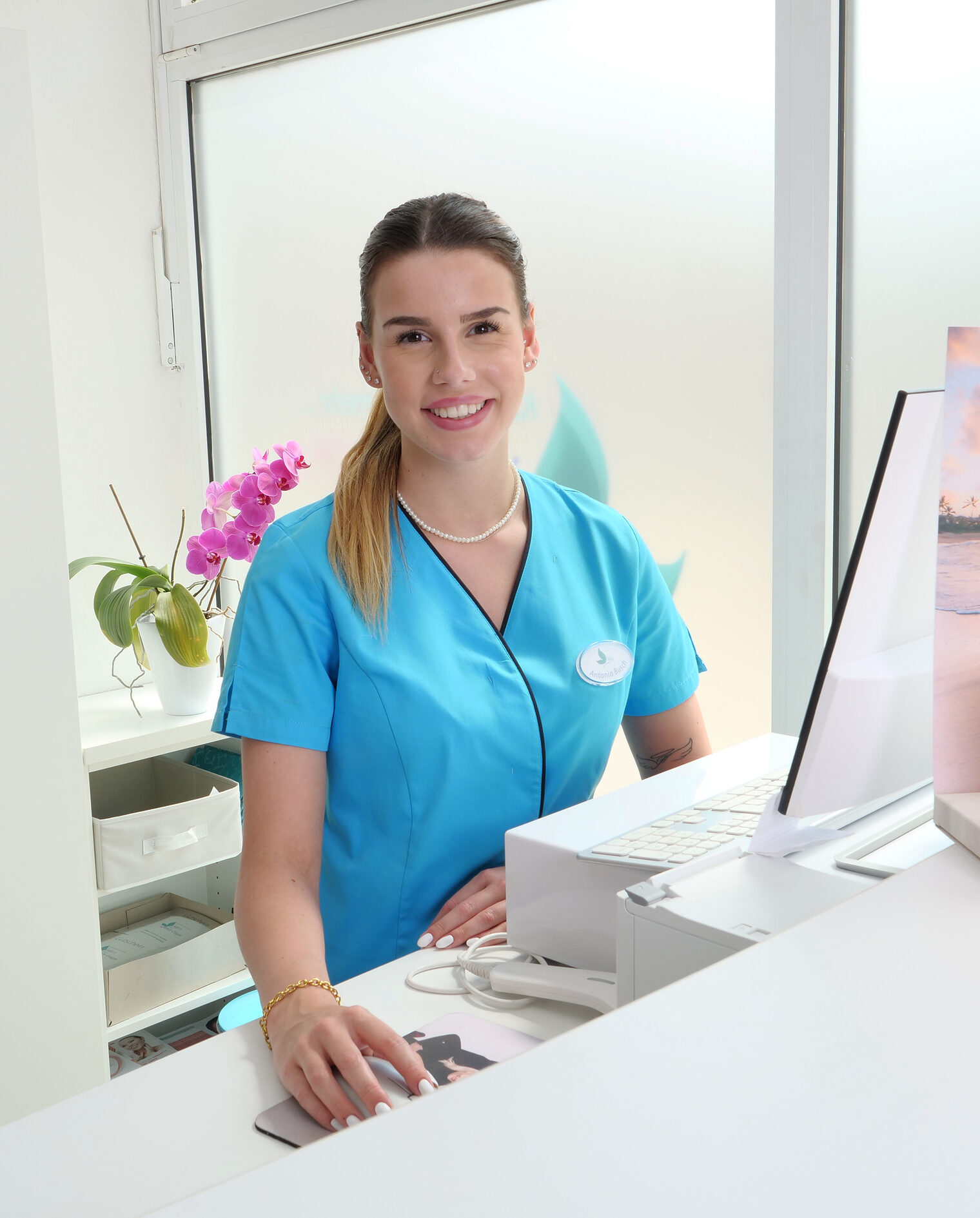 Frau in knallblauem T-Shirt am Tresen an einem Rechner, lächelt  Frau in knallblauem T-Shirt am Tresen an einem Rechner, lächelt in die Kamera