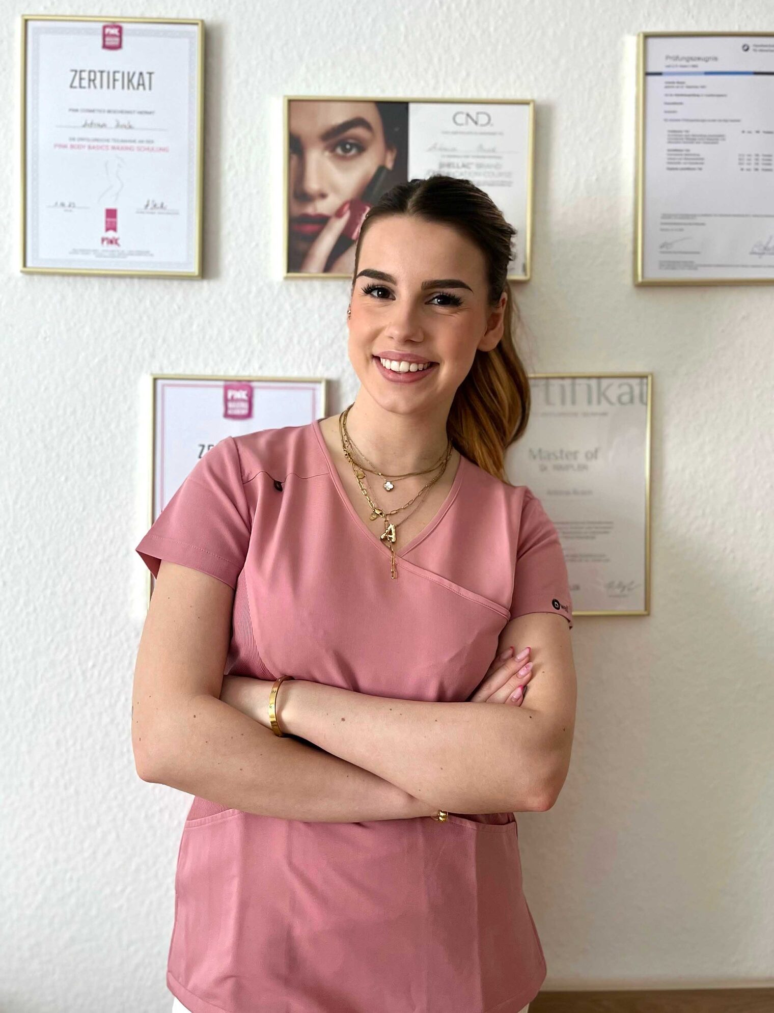 Frau vor Wand mit Zertifikaten in Rahmen, verschränkte Arme, lächelt Frau steht vor Wand mit Zertifikaten in Rahmen, verschränkte Arme, lächelt