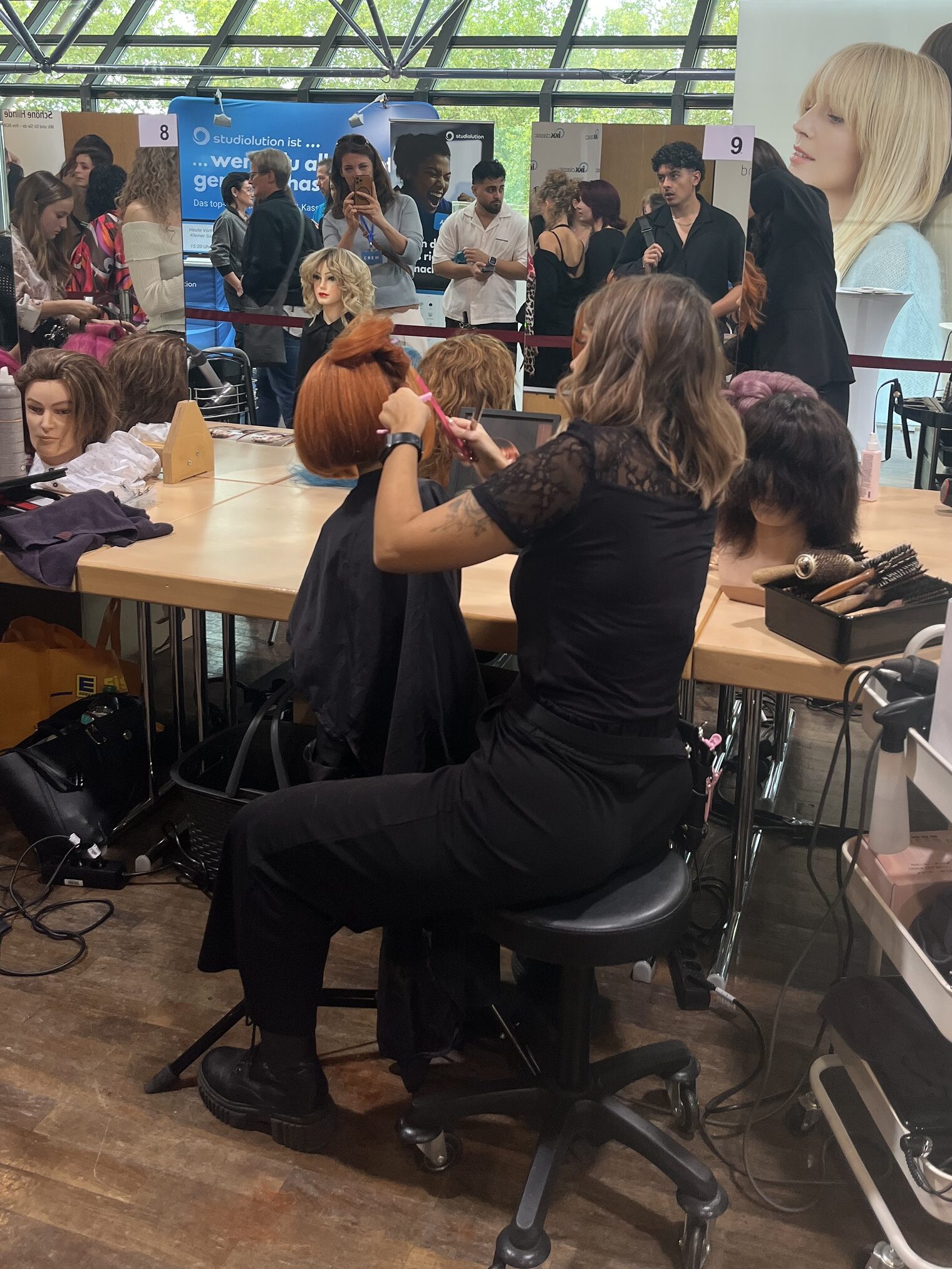 DMH 2025 | Kammersieger Junge Friseurin von hinten bei der Arbeit an einem Frisierkopf fotografiert. Im Hintergrund Wettbewerb mit Publikum und anderen Teilnehmerinnen.