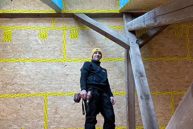 DMH 2025 | Kammersieger Zimmerer auf Baustelle unterm Dach, blickt nach unten in Kamera