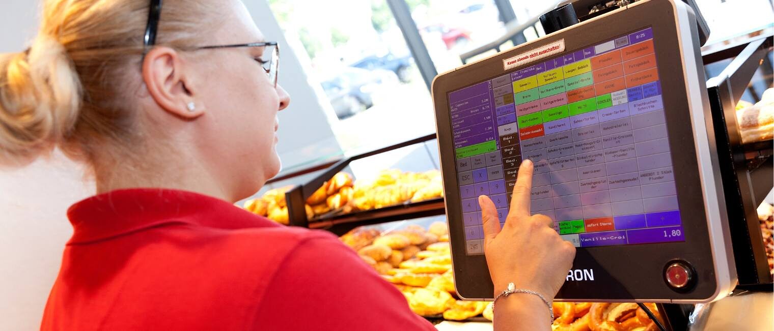 Eine Bäckereifachverkäuferin tippt hinter der Bäckertheke etwas auf einem Display der Kasse ein, trägt rotes T-Shirt und Zopf
