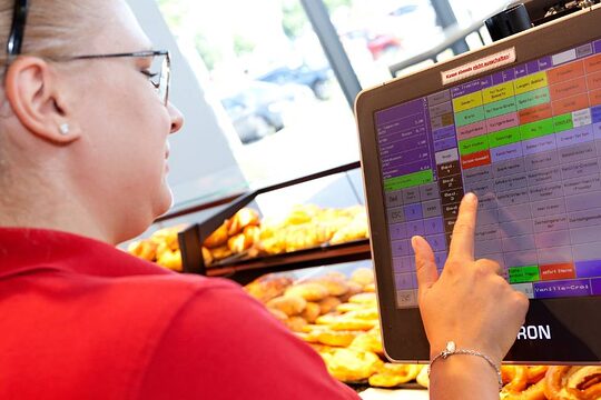 Eine Bäckereifachverkäuferin tippt hinter der Bäckertheke etwas auf einem Display der Kasse ein, trägt rotes T-Shirt und Zopf