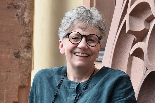 Frau mit kurzen grauen Haaren, schwarzer runder Brille und türkisem Oberteil lacht in die Kamera