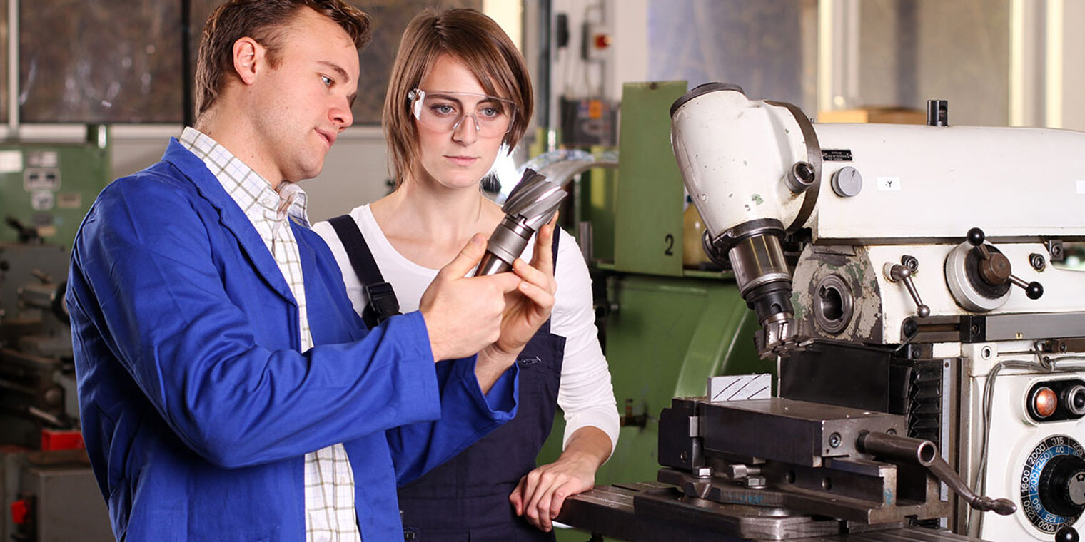 Ausbilder zeigt Azubine ein Werkstück, Sie trägt eine Schutzbrille, sie stehen in einer Werkstatt